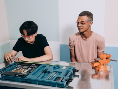 Dois jovens usando óculos de proteção trabalham juntos em um projeto com ferramentas, em uma oficina de formação técnica.