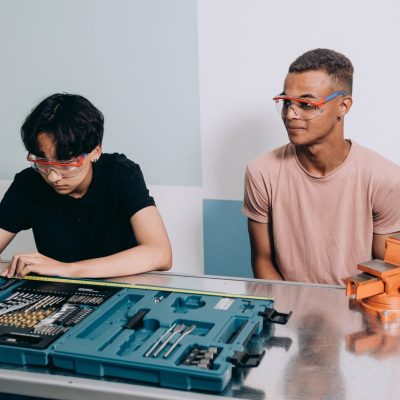 Dois jovens usando óculos de proteção trabalham juntos em um projeto com ferramentas, em uma oficina de formação técnica.