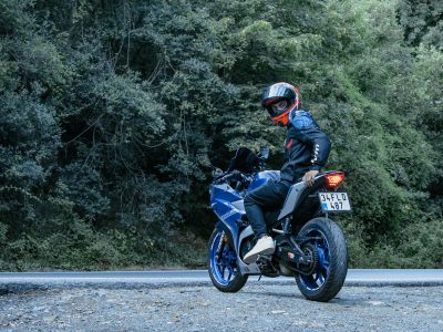 Motociclista usando capacete conduz uma motocicleta Yamaha azul por uma estrada cercada de vegetação.