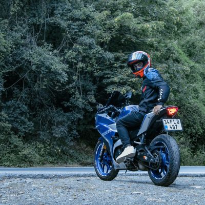 Motociclista usando capacete conduz uma motocicleta Yamaha azul por uma estrada cercada de vegetação.