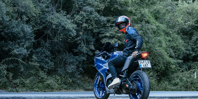 Motociclista usando capacete conduz uma motocicleta Yamaha azul por uma estrada cercada de vegetação.