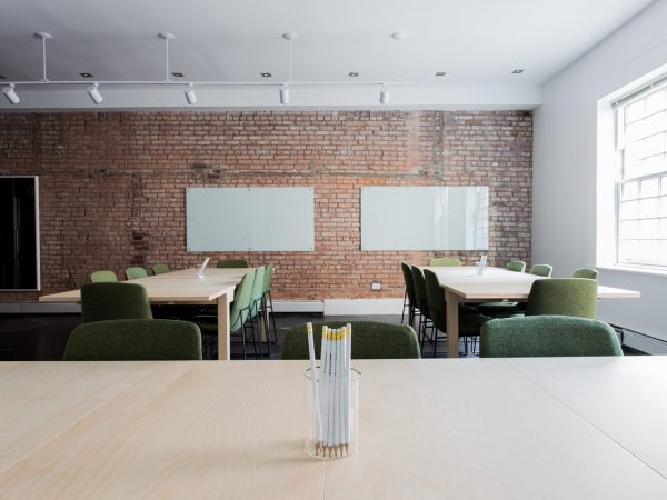 Sala de aula vazia com mesas, cadeiras e paredes de tijolos aparentes.