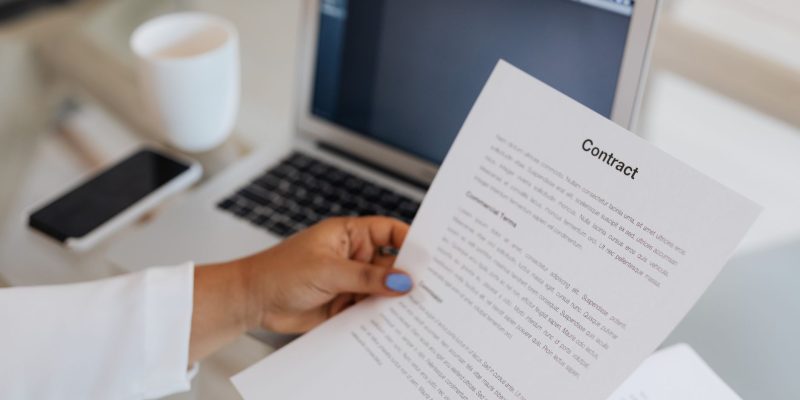Pessoa segura um documento contratual ao lado de um computador portátil durante uma sessão de trabalho.